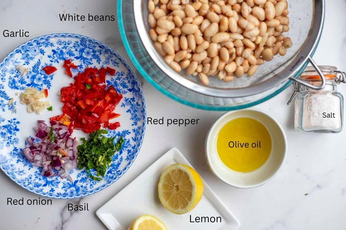 white beans drained in a strainer, jar of salt, bowl of oil, sliced lemon, and a plate of chopped vegetables and herbs.