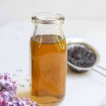 Lilac syrup in a bottle with strained flowers in the background and fresh flowers in the front.