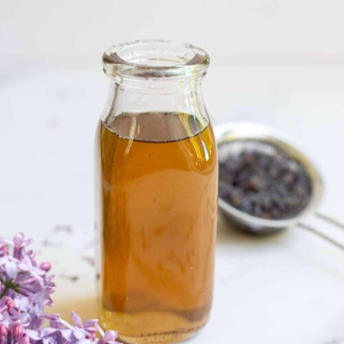 Lilac syrup in a bottle with strained flowers in the background and fresh flowers in the front.