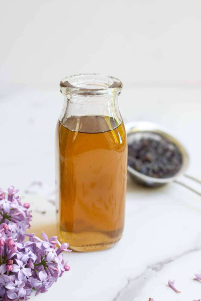 Lilac syrup in a bottle with strained flowers in the background and fresh flowers in the front.