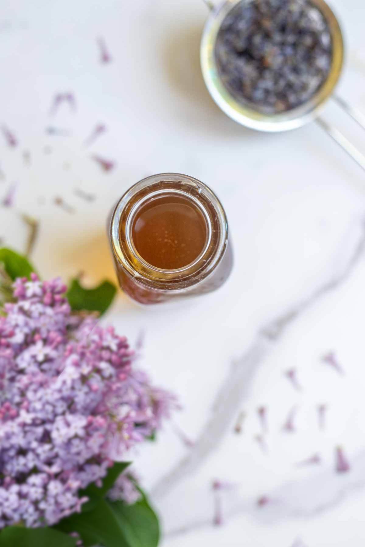 overhead photo of lilac syrup in a jar with fresh lilac flowers and steeped strain flowers surrounding the jar.