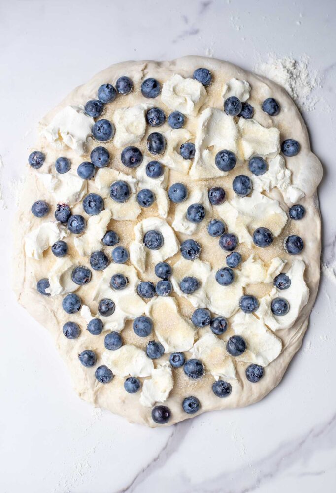 Sourdough bread dough patted out into a large circle and topped with cream cheese, blueberries and sugar.