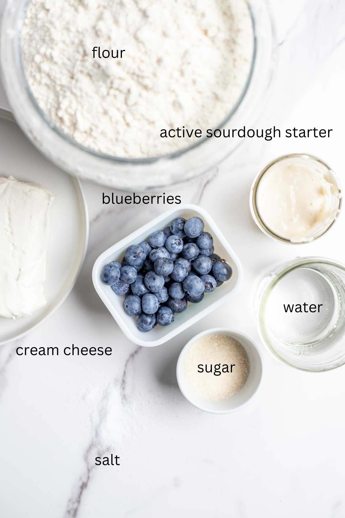 containers of flour, sourdough starter, water sugar, fresh blueberries and a plate with a block of cream cheese on a marble countertop.