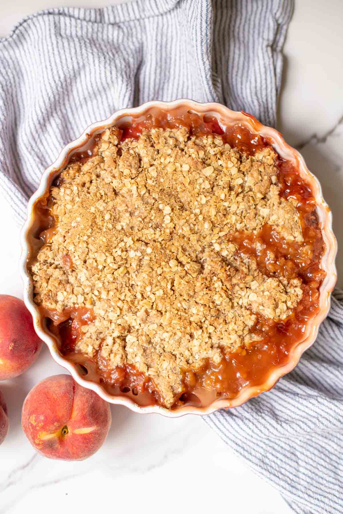 Overhead photo of sourdough peach crisp in a deep pie plate on a towel next to peaches.
