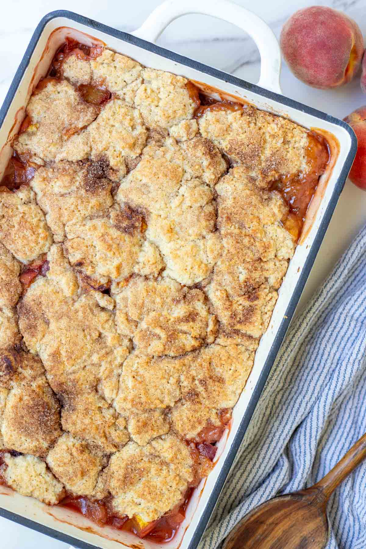 Sourdough peach cobbler in a white baking dish surrounded by peaches and a towel.