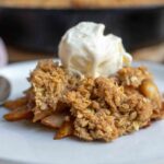 Pear crisp topped with a scoop of ice cream on top on a plate with a cast irons skillet of more crisp in the background.