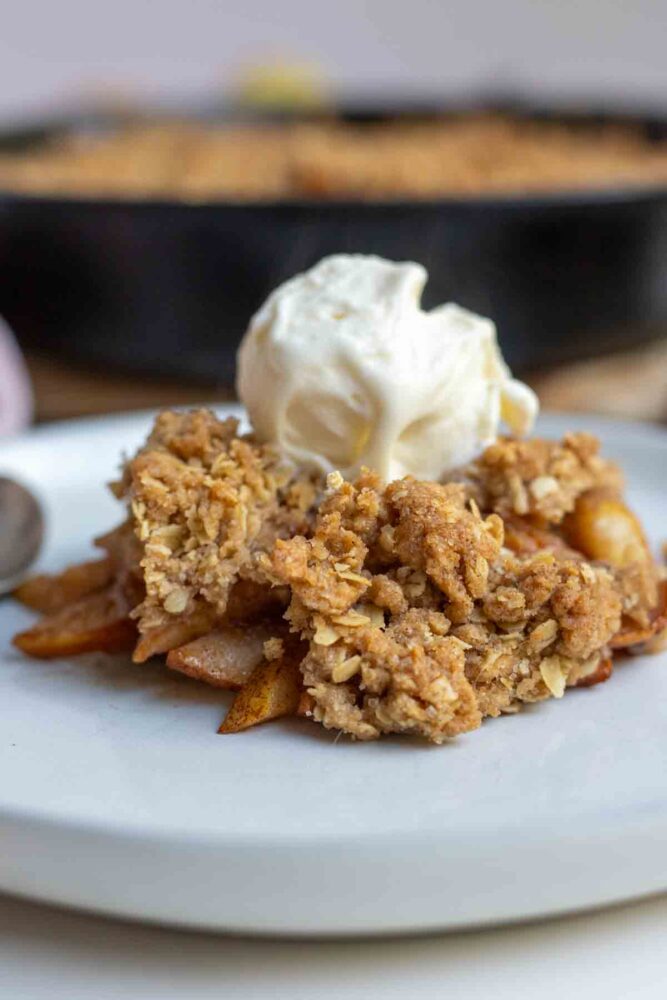 Pear crisp topped with a scoop of ice cream on top on a plate with a cast irons skillet of more crisp in the background.
