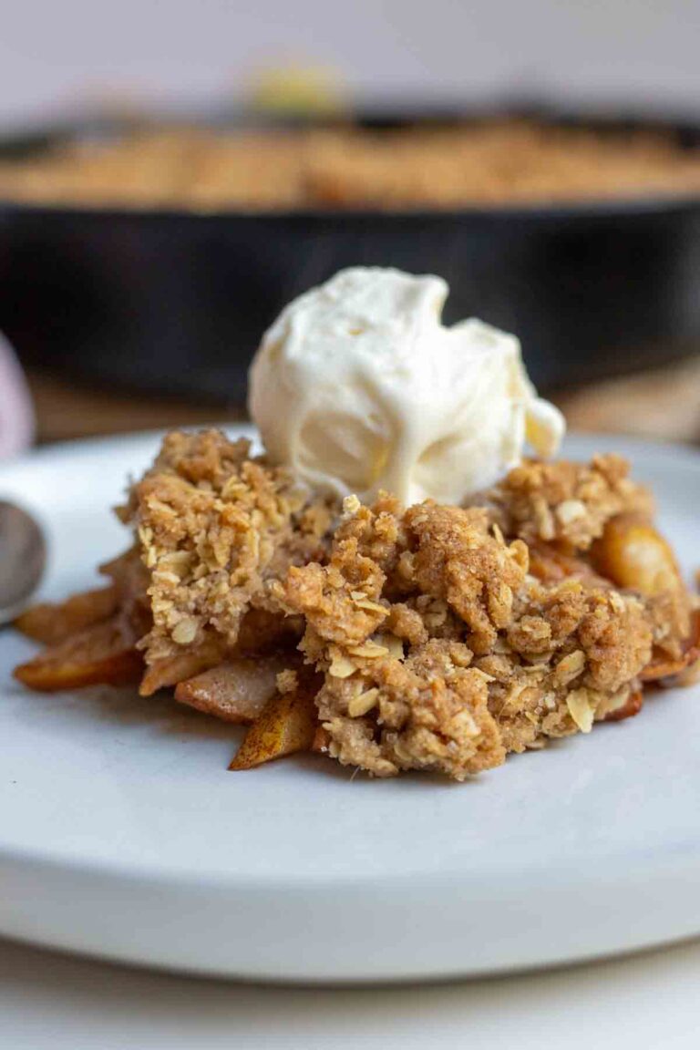 Pear crisp topped with a scoop of ice cream on top on a plate with a cast irons skillet of more crisp in the background.