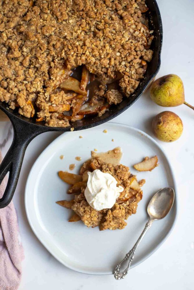 Overhead photo of pear crisp topped with ice cream on a plate with a spoon and a skillet of pear crisp in the background.