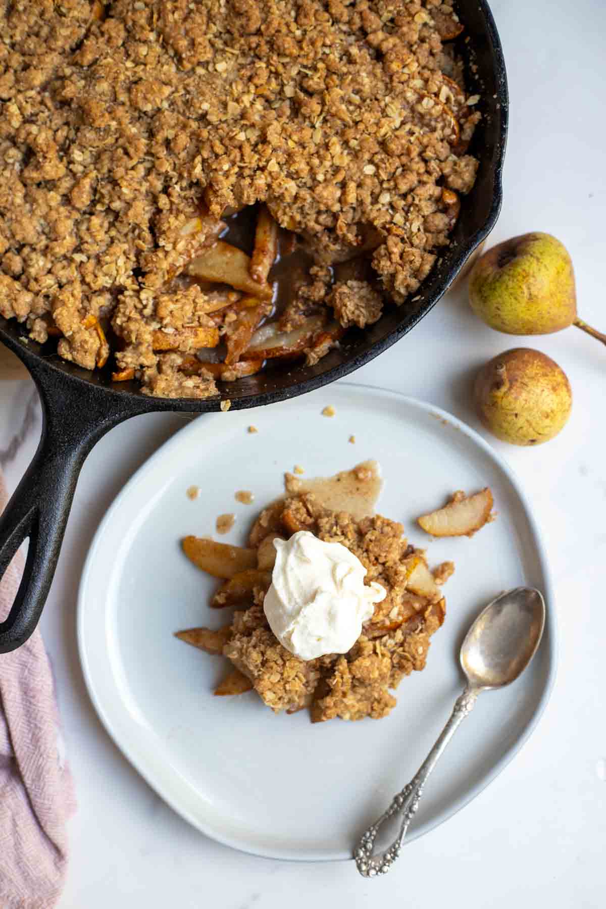 Overhead photo of pear crisp topped with ice cream on a plate with a spoon and a skillet of pear crisp in the background.