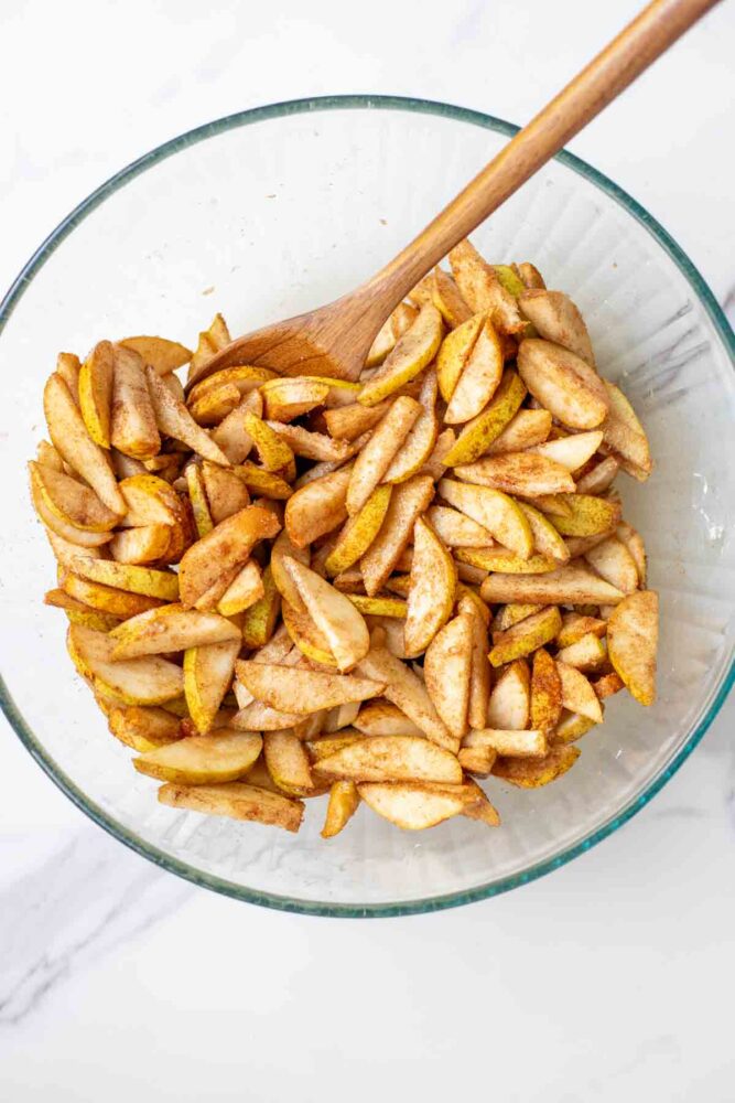 Sliced pears mixed with spices, brown sugar and cornstarch in a glass bowl with a wooden spoon.