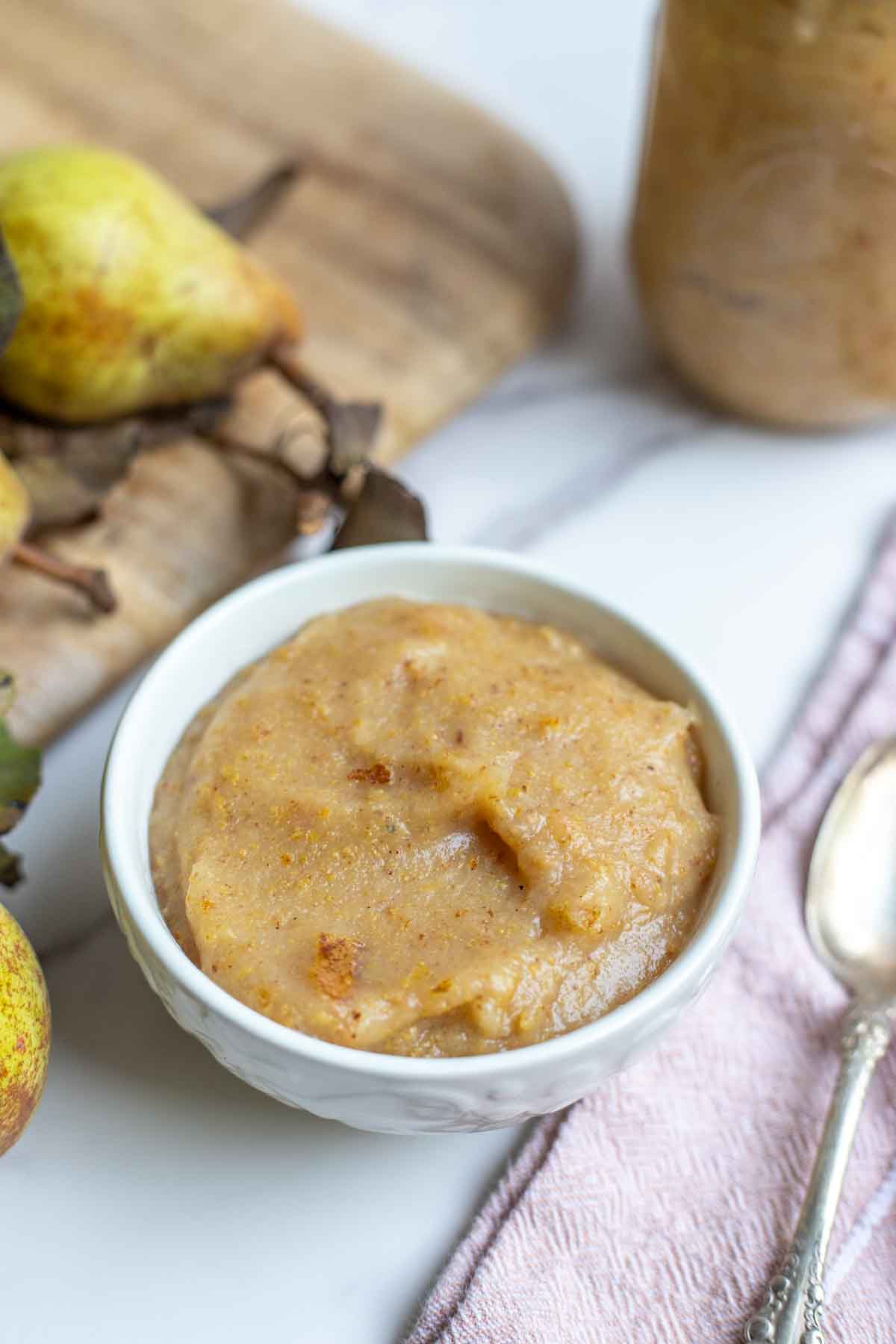 Homemade pear sauce in a bowl. A pink napkin with a spoon is to the right of the bowl, and cutting board with more pears are behind the bowl.