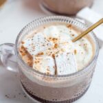 Close up picture of pumpkin spice hot chocolate topped with whipped cream, marshmallows, and pumpkin spice with a gold spoon. More hot chocolate in th background.