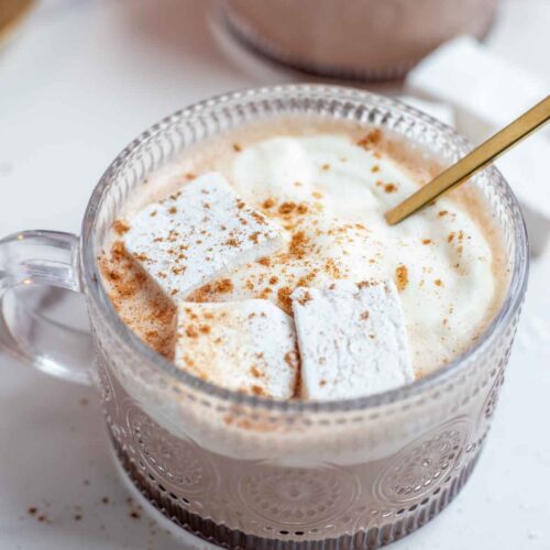 Close up picture of pumpkin spice hot chocolate topped with whipped cream, marshmallows, and pumpkin spice with a gold spoon. More hot chocolate in th background.
