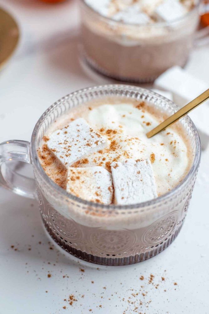 Close up picture of pumpkin spice hot chocolate topped with whipped cream, marshmallows, and pumpkin spice with a gold spoon. More hot chocolate in th background.