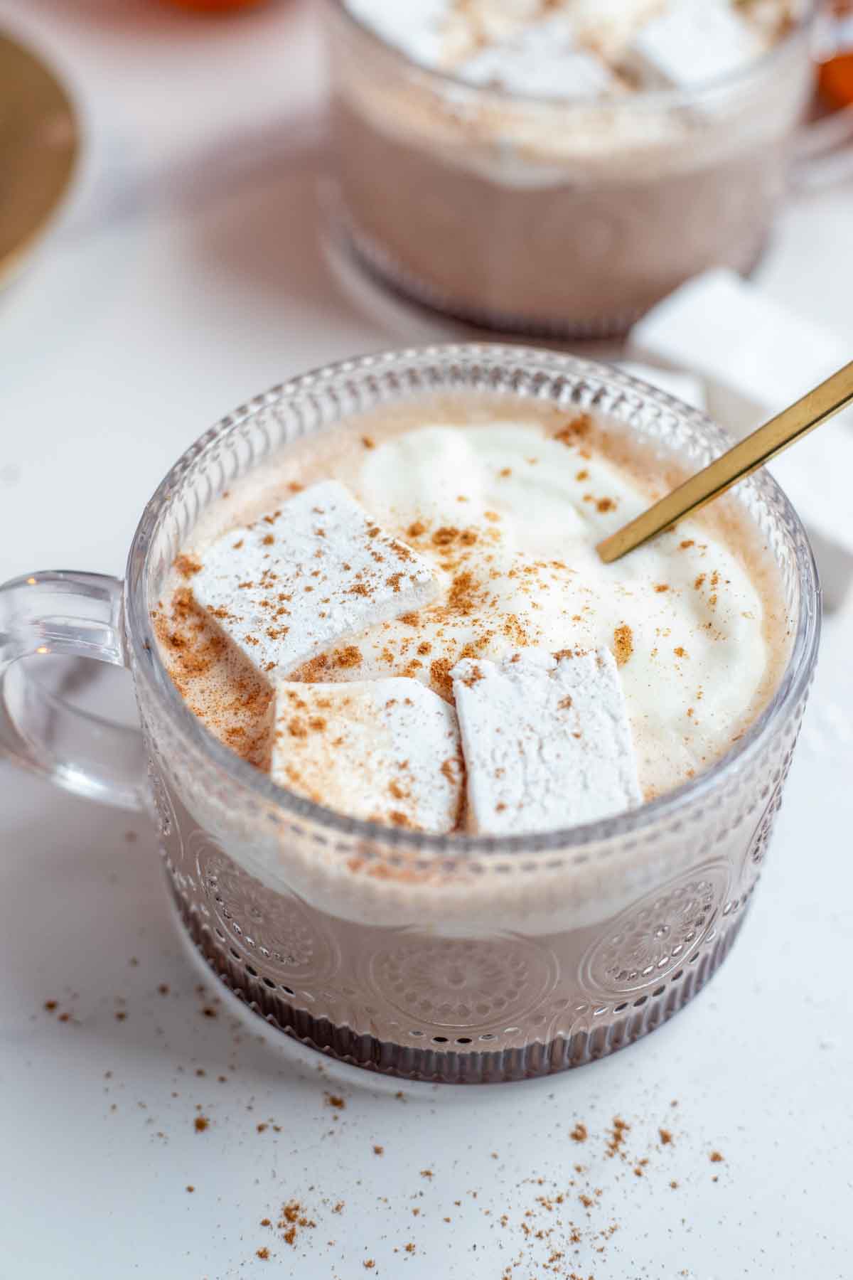 Close up picture of pumpkin spice hot chocolate topped with whipped cream, marshmallows, and pumpkin spice with a gold spoon. More hot chocolate in th background.
