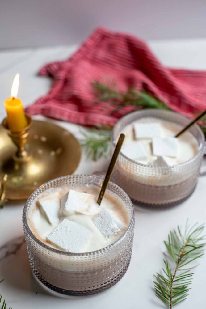 Two mugs of creamy hot chocolate topped with homemade marshmallows and whipped cream with brass spoons in each mug. a candle, towel and greenery are in the background.