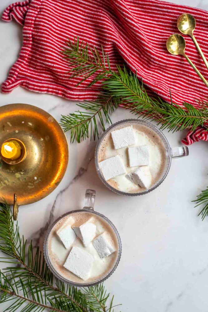 Two glass mugs of healthy hot chocolate topped with homemade marshmallows. Greenery, a candle, red towel with brass spoons surrounding the mugs.