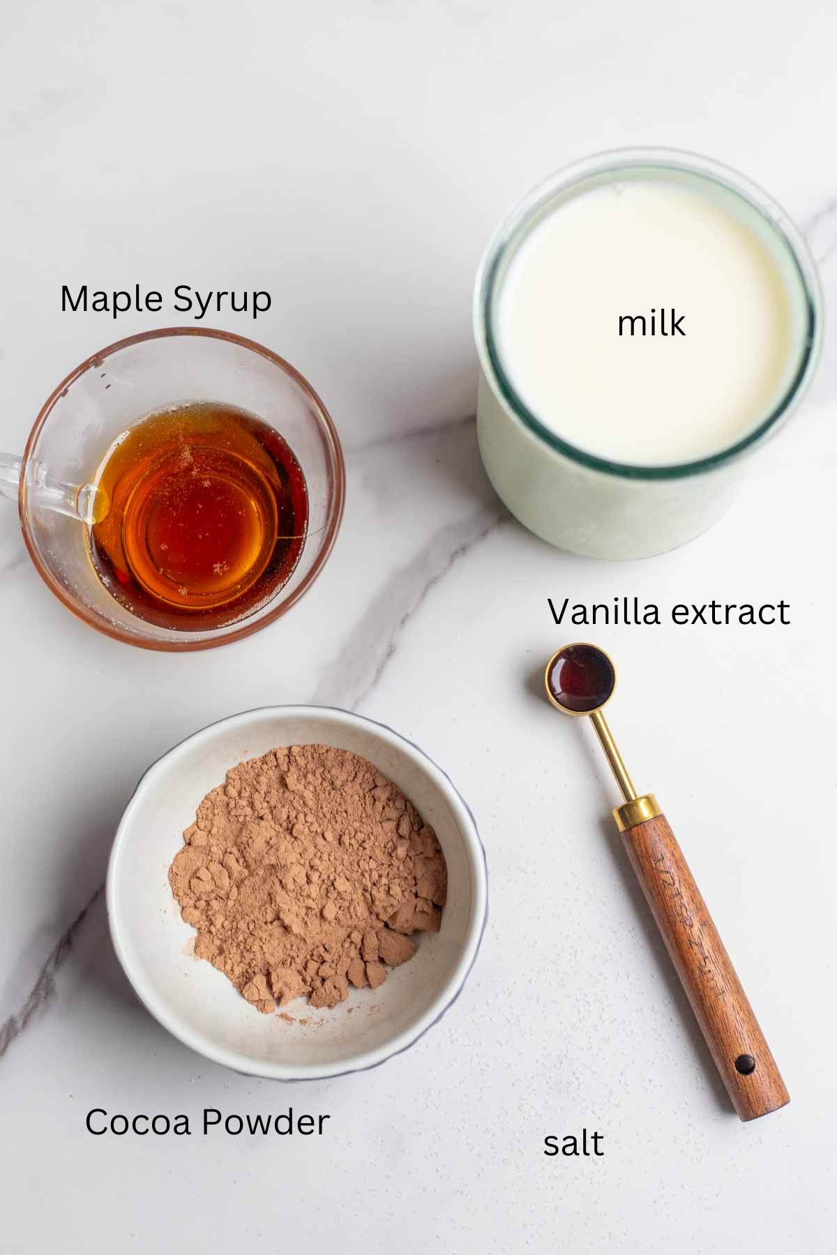 small bowls of cocoa powder and maple syrup, a glass of milk and a measuring spoon of vanilla on a countertop.
