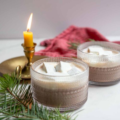 Two mugs of healthy hot chocolate topped with homemade marshmallows and whipped cream. The mugs are surrounded by greenery and a candle.