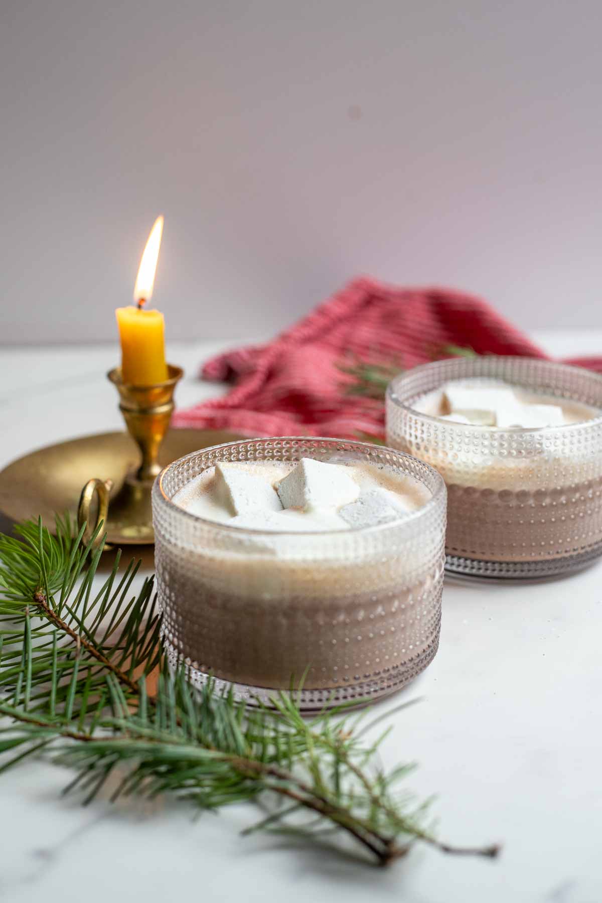 Two mugs of healthy hot chocolate topped with homemade marshmallows and whipped cream. The mugs are surrounded by greenery and a candle.
