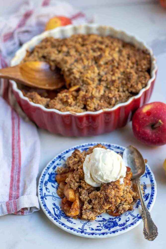 Sourdough apple crisp on a plate and topped with ice cream. More crisp is in the background in a red baking dish.