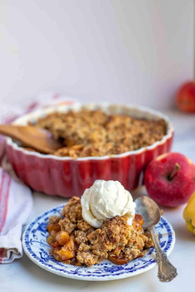 Sourdough apple crisp topped with ice cream on a plate with a red baking dish full of crisp and apples in the background.
