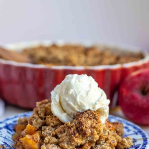 Sourdough Apple Crisp topped with ice cream on a blender and white plate with a red baking dish of crisp in the background.
