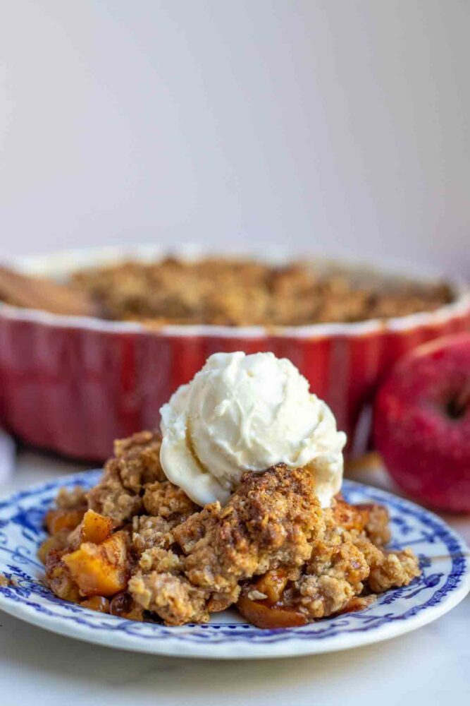 Sourdough Apple Crisp topped with ice cream on a blender and white plate with a red baking dish of crisp in the background.