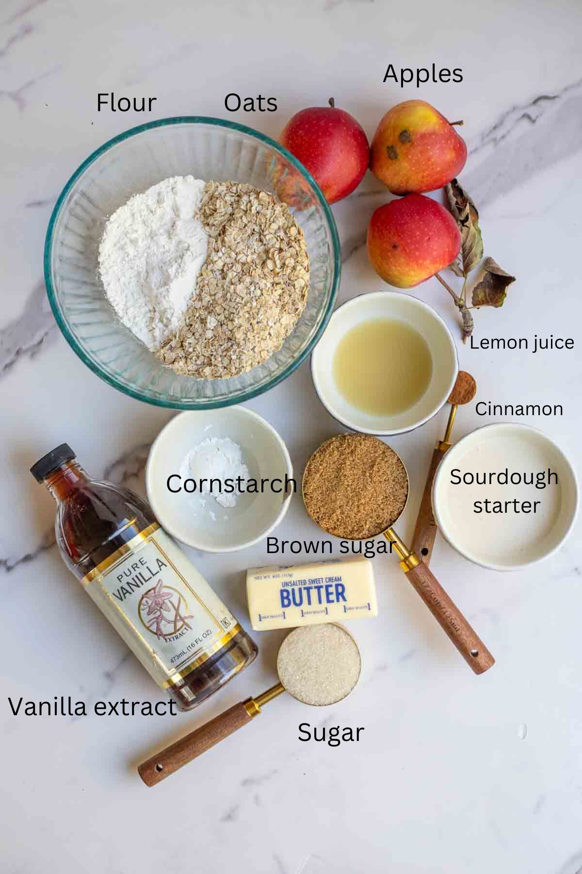 Apples, oats, flour, sourdough starter, cinnamon and other ingredients on a marble countertop.
