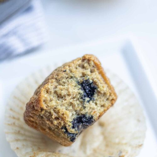 Whole wheat sourdough blueberry muffin with fresh milled flour cut in half on a muffin wrapper on a white plate.