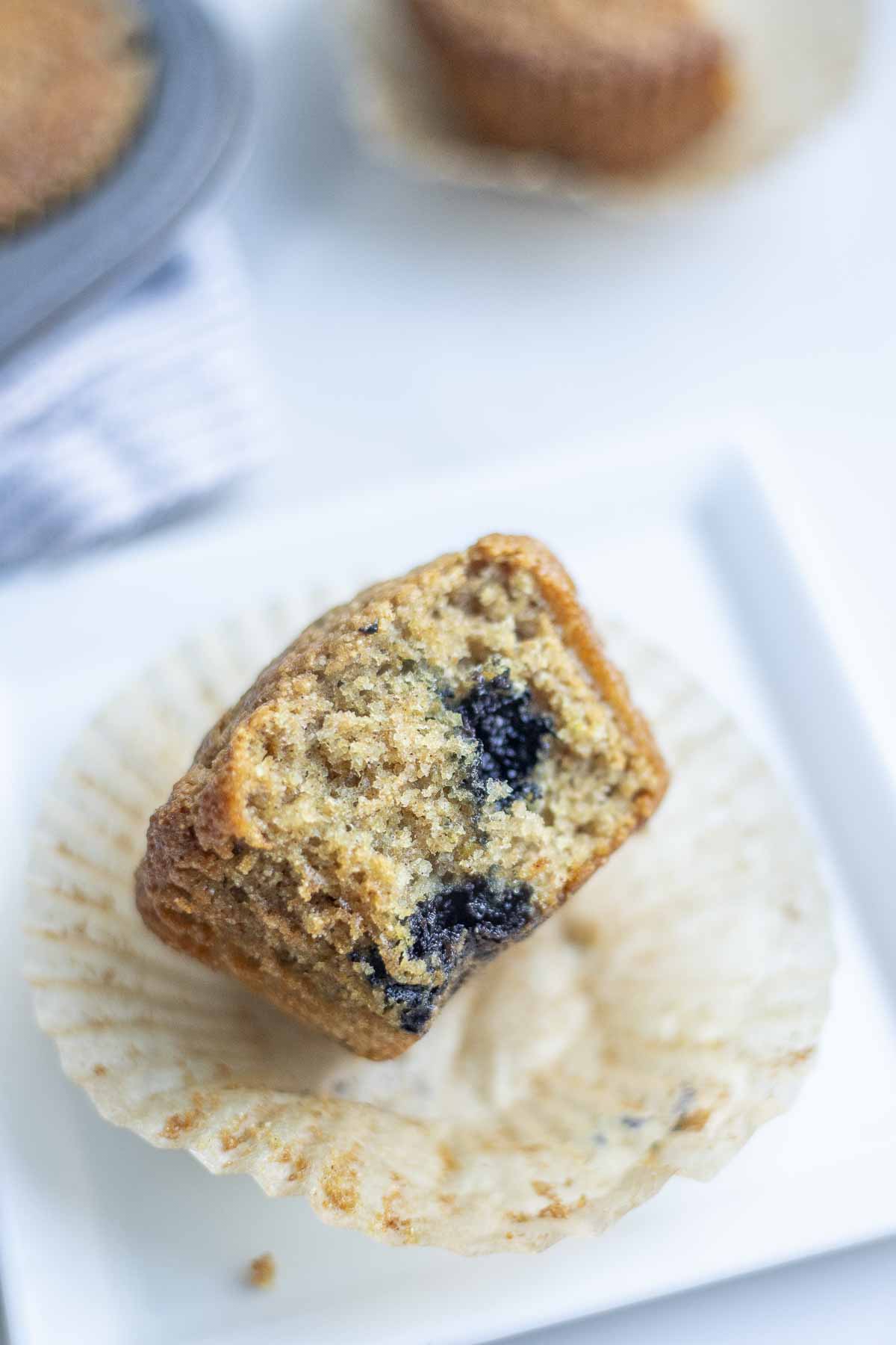 Whole wheat sourdough blueberry muffin with fresh milled flour cut in half on a muffin wrapper on a white plate.
