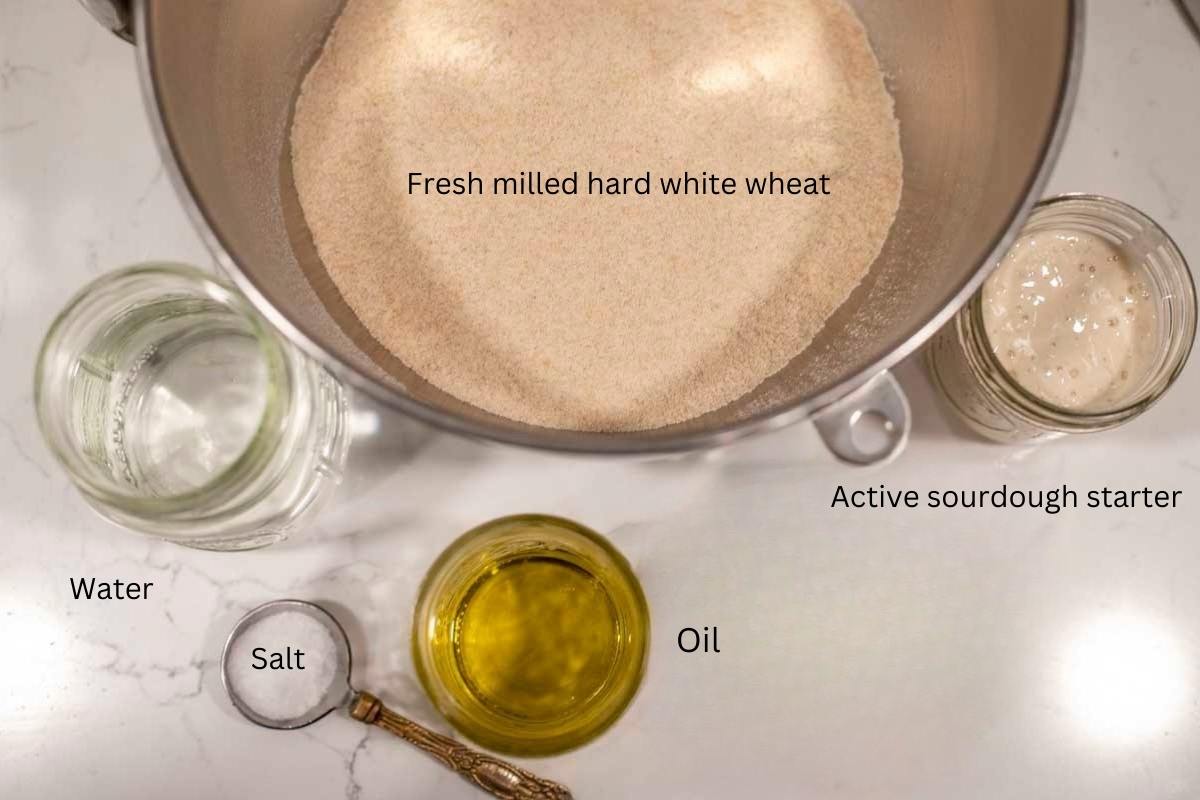 fresh milled flour in a bowl. oil, water, active starter in jars, and a spoon of salt on a countertop.