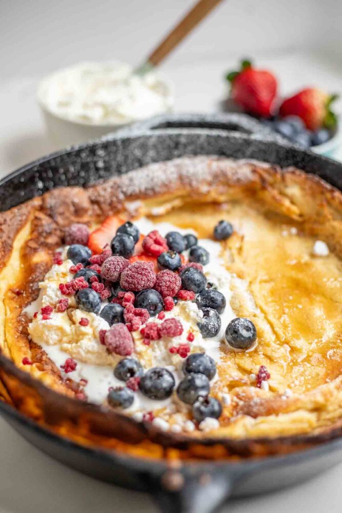 dutch baby pancake topped with whipped cream, berries, and powdered sugar. A Bowl of whipped cream and strawberries in the background.