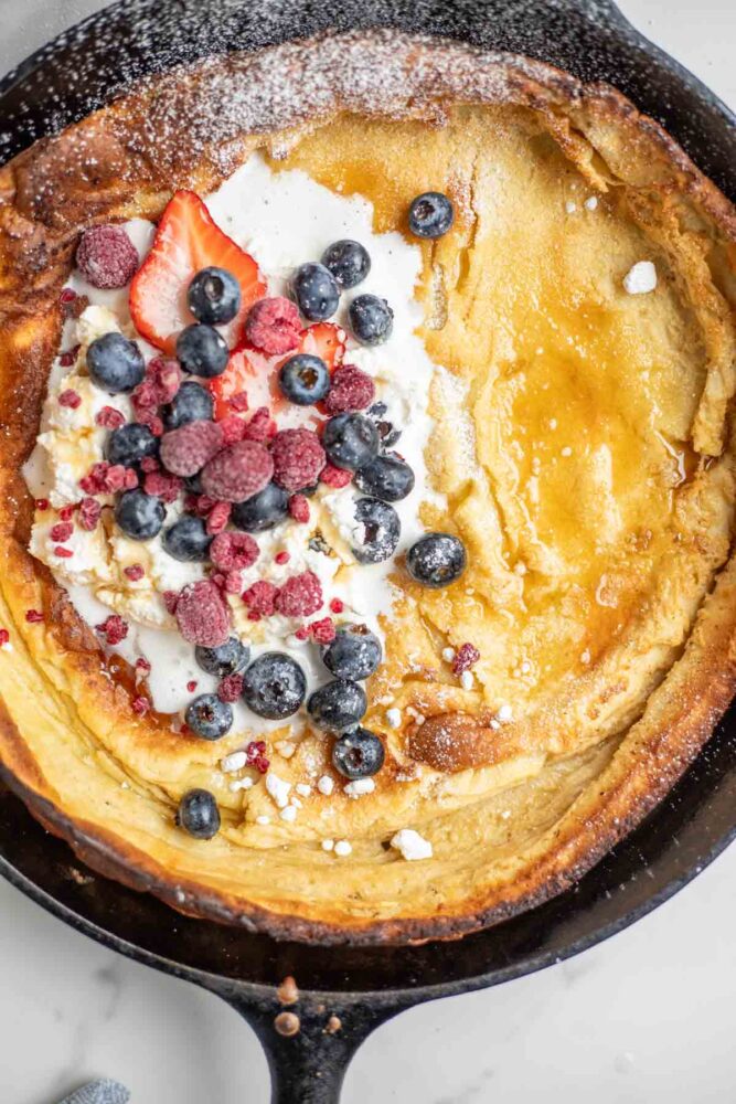 Overhead close up photo of a dutch baby pancake with whipped cream, berries, powdered sugar and maple syrup in a cast iron skillet.