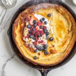 Dutch baby pancake topped with whipped cream, blueberries, strawberries, raspberries, and powdered sugar in a cast iron skillet. The skillet is surrounded by blueberries and a sifter of powdered sugar.