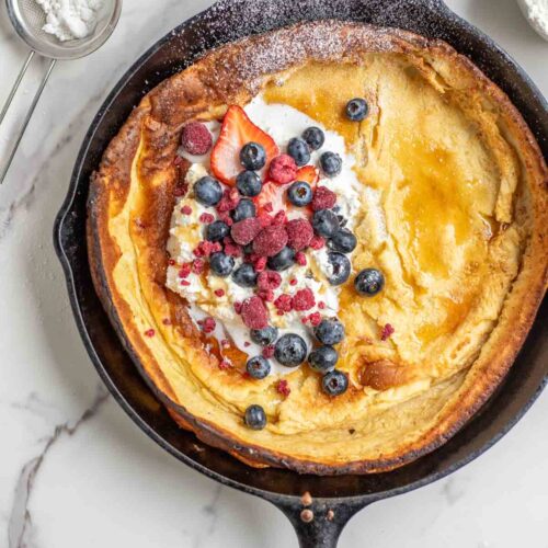 Dutch baby pancake topped with whipped cream, blueberries, strawberries, raspberries, and powdered sugar in a cast iron skillet. The skillet is surrounded by blueberries and a sifter of powdered sugar.