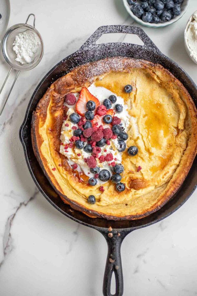 Dutch baby pancake topped with whipped cream, blueberries, strawberries, raspberries, and powdered sugar in a cast iron skillet. The skillet is surrounded by blueberries and a sifter of powdered sugar.