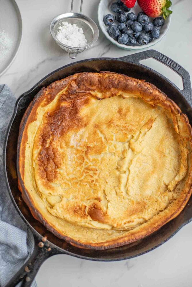 Baked dutch pancake in a skillet with blueberries and strawberries in a bowl, and powdered sugar in a stainer.