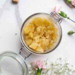 Overhead photo of honey sugar scrub in a glass container with a flip top. The jar is surrounded by flowers.