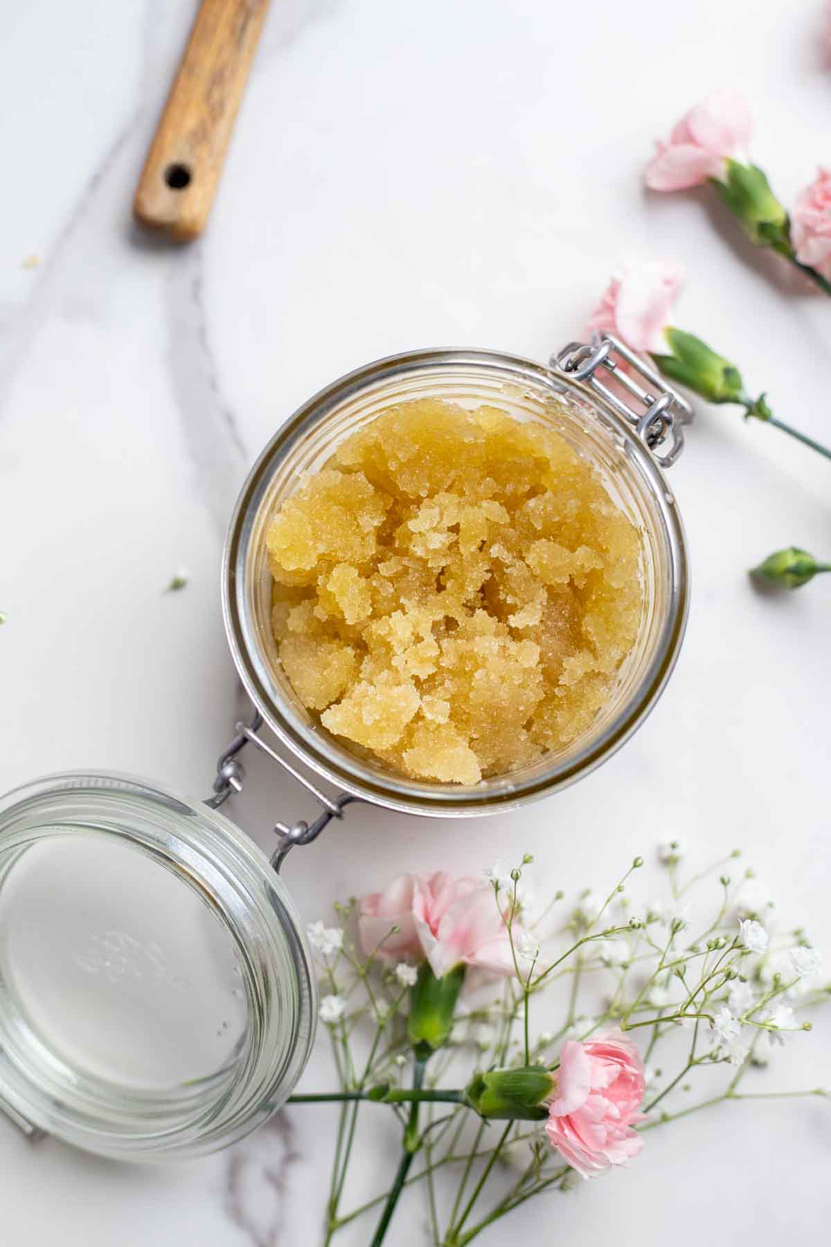 Overhead photo of honey sugar scrub in a glass container with a flip top. The jar is surrounded by flowers.