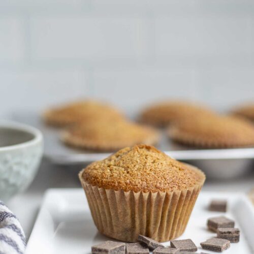 Whole wheat sourdough chocolate chip muffin on a plate surrounded by chocolate chips with more muffins in the background.