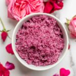 Overhead photo of a bowl of rose sugar surrounded by roses on a marble countertop.