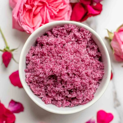 Overhead photo of a bowl of rose sugar surrounded by roses on a marble countertop.