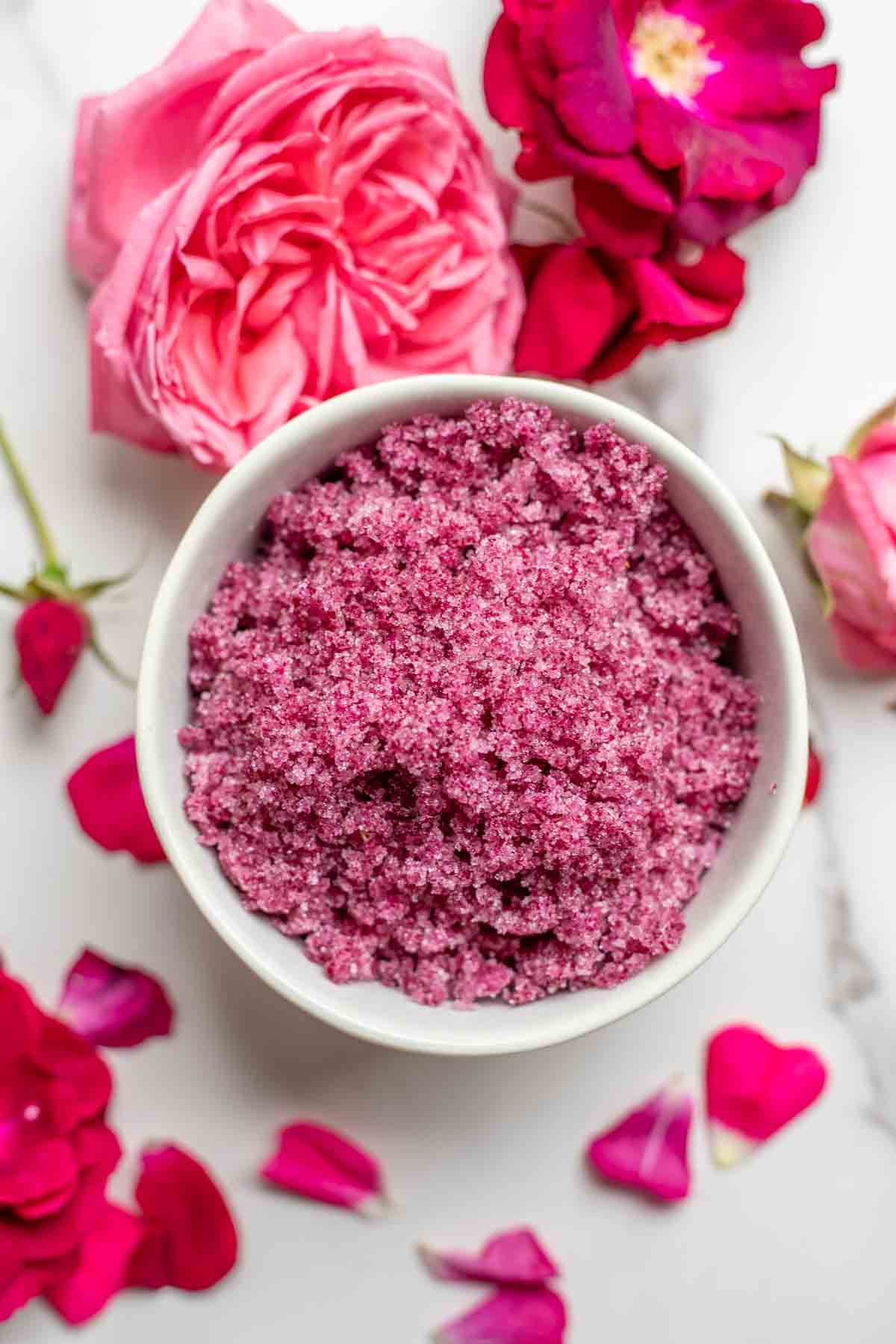 Overhead photo of a bowl of rose sugar surrounded by roses on a marble countertop.