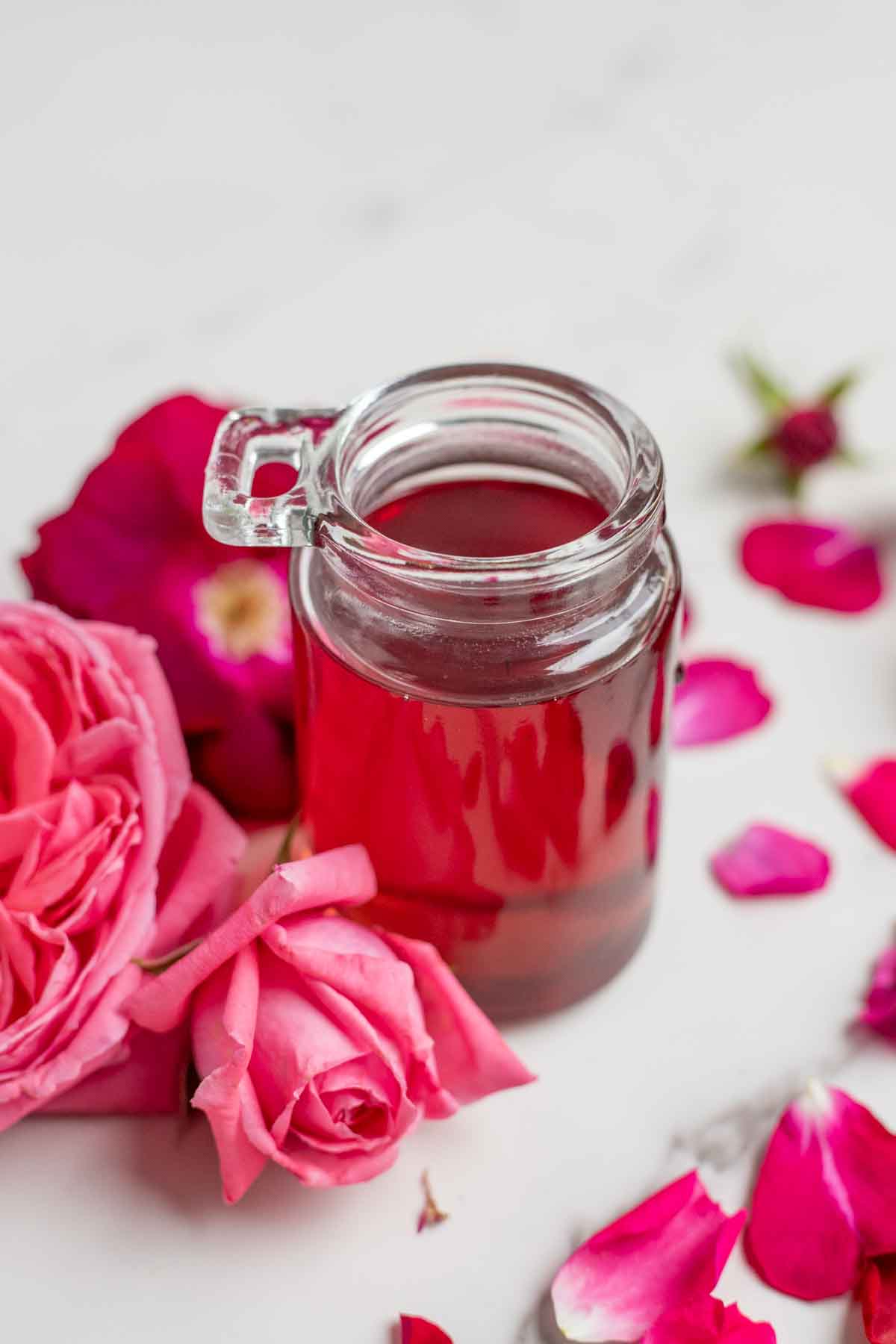 Rose syrup in a small glass jar surrounded by pink and red roses.