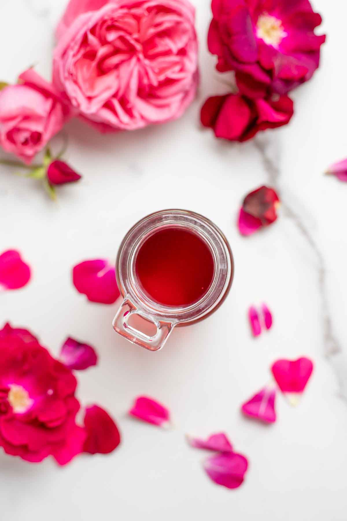 Overhead photo of rose syrup in a glass jar surrounded by roses and rose petals.