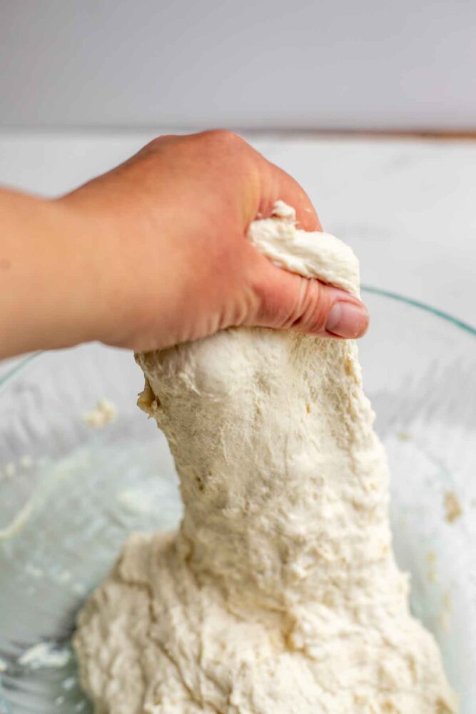 A hand grabbing dough and pulling it up out of a bowl to complete a stretch and fold.