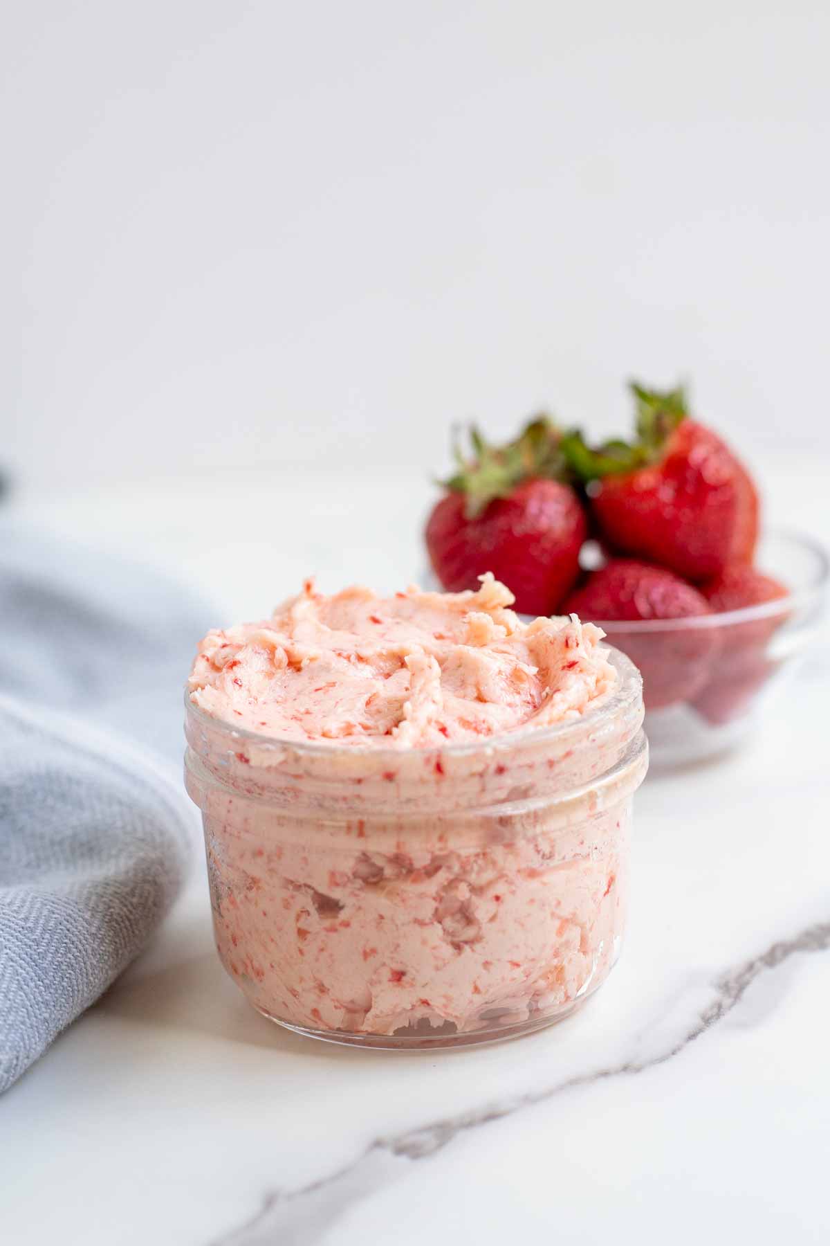 Strawberry butter in a glass jar with strawberries and a blue towel in the background.
