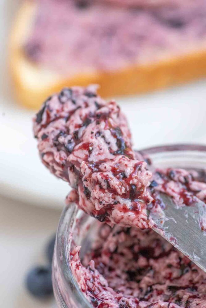 Knife full of blueberry butter out of a jar with a piece of toast in the background.